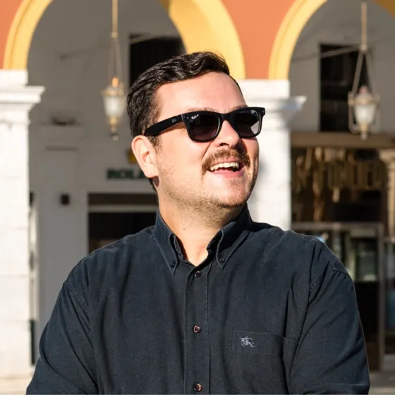 Victor Adbib, en chemise sombre et lunettes de soleil, sourit sur une place ensoleillée